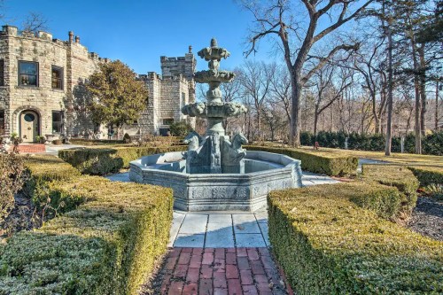 A decorative stone fountain surrounded by neatly trimmed hedges in a sunny garden setting.
