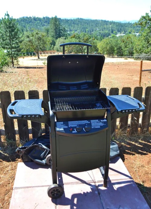 A black gas grill with an open lid and side tables, set on a patio with a scenic outdoor background.