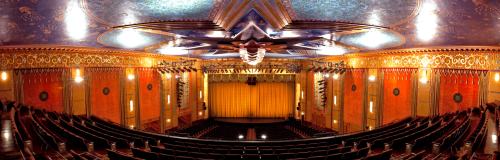A grand theater interior with ornate ceiling, vibrant colors, and a large stage curtain, viewed from the balcony.
