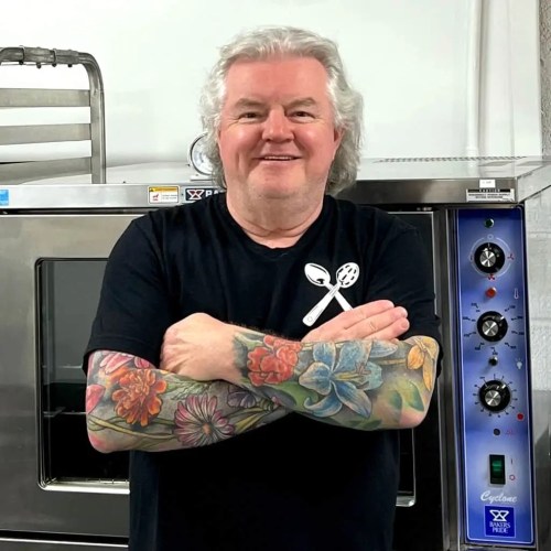 A smiling man with colorful floral tattoos stands in a kitchen, arms crossed, next to a large oven.