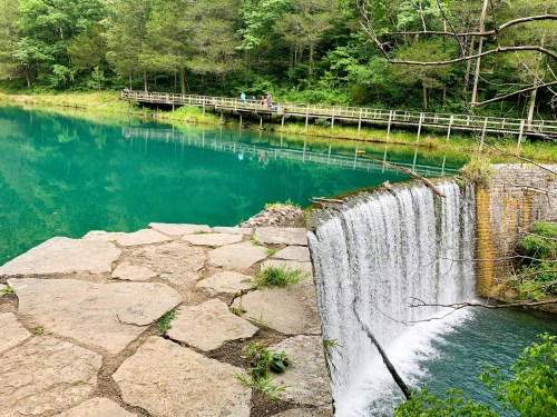 A serene scene featuring a waterfall cascading into a turquoise lake, surrounded by lush greenery and a wooden walkway.