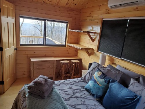 Cozy wooden cabin interior with a bed, shelves, and a scenic window view of nature.