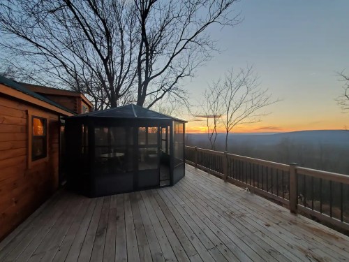 A wooden deck with a screened gazebo overlooks a sunset and distant hills, surrounded by bare trees.