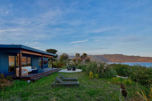 A modern blue house with a deck overlooks a lush garden and scenic mountains by the water under a clear sky.