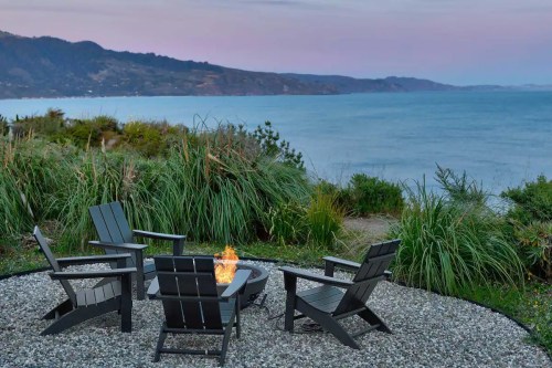 A cozy fire pit surrounded by chairs, overlooking a serene ocean view and lush greenery at sunset.