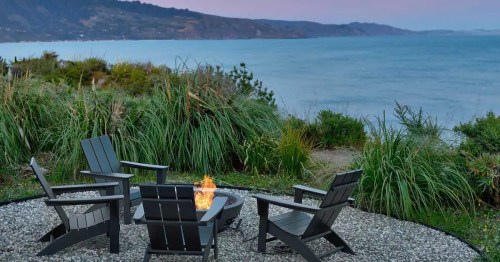 A cozy fire pit surrounded by black chairs, overlooking a serene lake and lush greenery at dusk.