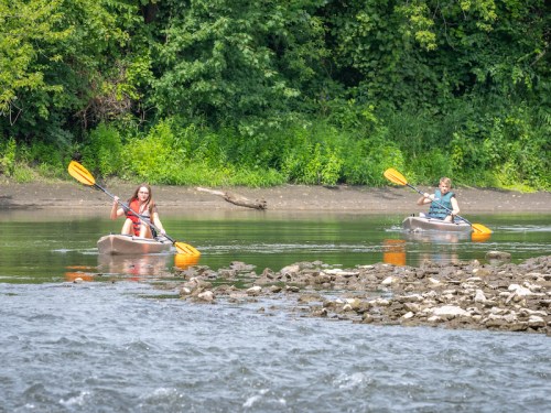 Two people kayaking on a calm river, surrounded by lush greenery and rocky riverbank.