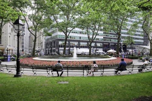 A park scene with a circular fountain surrounded by flowers, benches, and tall buildings in the background.