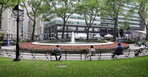 A park scene with benches, a fountain, and people sitting among flower beds and trees in an urban setting.