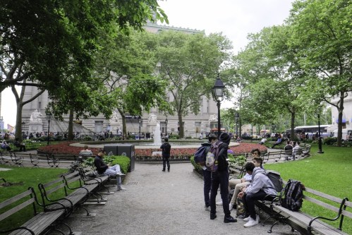 A park scene with benches, greenery, and people socializing near flower beds and a historic building in the background.