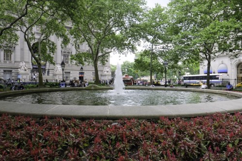 A circular fountain surrounded by greenery and colorful flowers in a park setting, with buildings in the background.