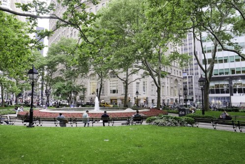 A park scene with green grass, benches, a fountain, and trees, surrounded by buildings and people enjoying the space.