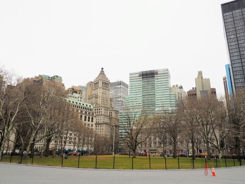 A cityscape featuring a mix of historic and modern buildings, with bare trees and a grassy area in the foreground.