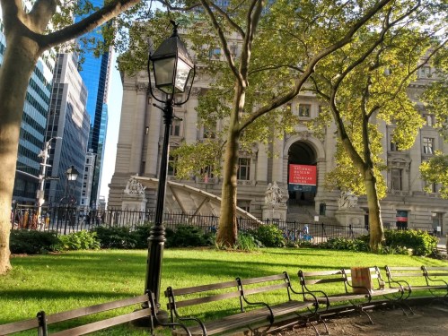 A park scene with benches, trees, and a historic building in the background, featuring a large banner.