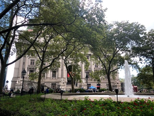 A historic building surrounded by trees and a fountain, with people sitting in a park area.