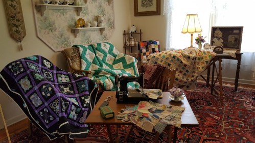A cozy room with quilts draped over furniture, a sewing machine on a table, and a warm, inviting atmosphere.