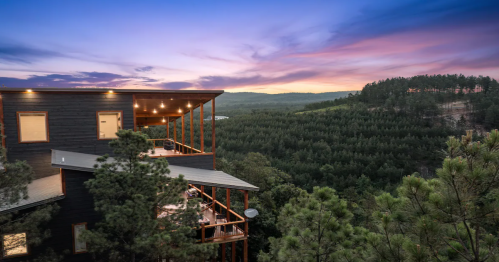 A modern cabin nestled in trees, overlooking a lush valley at sunset with vibrant skies.