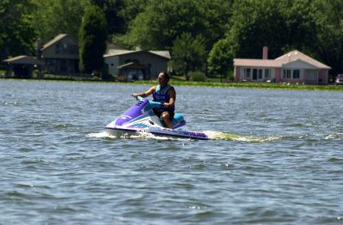 A person rides a jet ski on a lake, with houses and trees visible in the background on a sunny day.