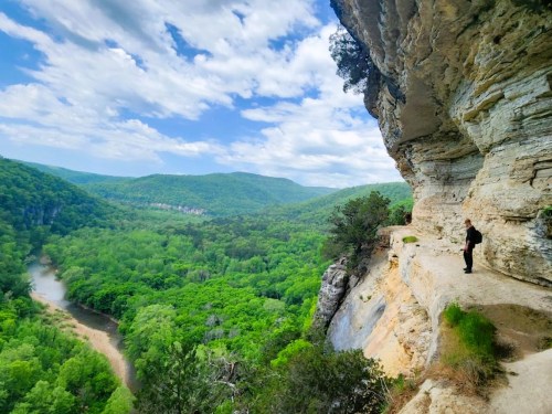 A person stands on a rocky ledge overlooking a lush green valley and river, under a bright blue sky with clouds.