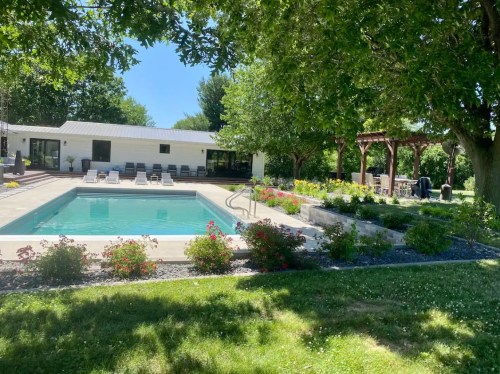 A sunny backyard with a pool, surrounded by greenery and flowers, featuring a patio and outdoor seating area.