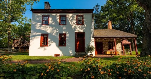 A charming two-story house with red trim, surrounded by greenery and yellow flowers under a clear blue sky.
