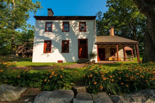 A two-story white house with red trim, surrounded by green grass and yellow flowers, under a clear blue sky.