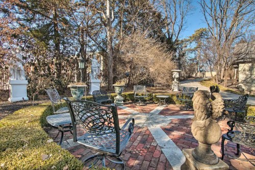 A serene garden with brick pathways, decorative statues, and wrought iron chairs surrounded by trees and greenery.