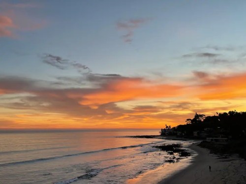 A serene beach at sunset, with vibrant orange and pink clouds reflecting on calm waters and a silhouette of a person walking.