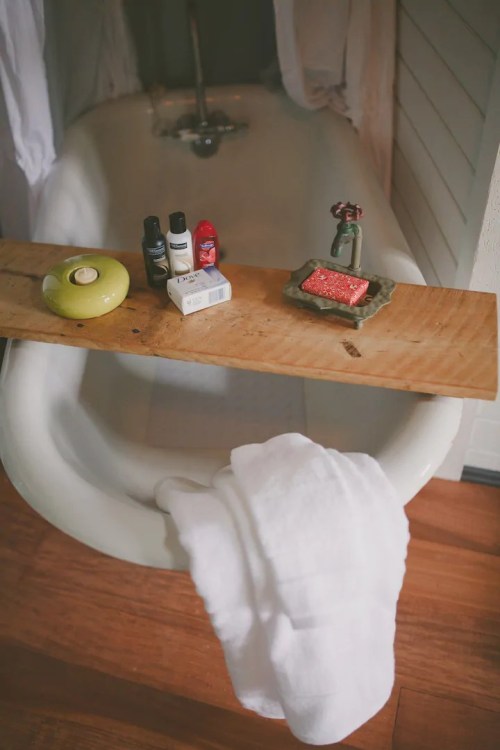A wooden tray over a bathtub holds toiletries, a soap bar, and a green dish, with a white towel draped nearby.