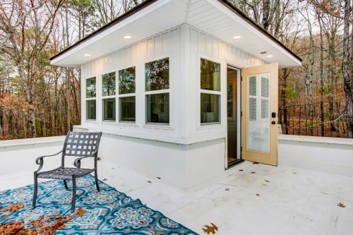 A cozy outdoor space with a chair, colorful rug, and large windows, surrounded by trees in a serene setting.