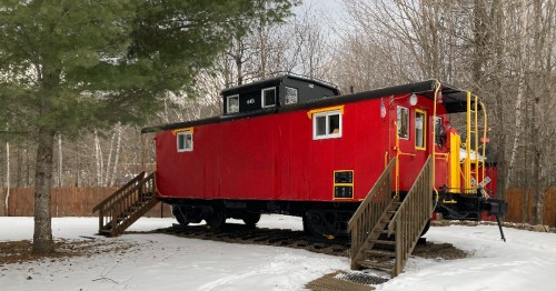 A red caboose on a snowy landscape, surrounded by trees and featuring a wooden staircase leading up to it.