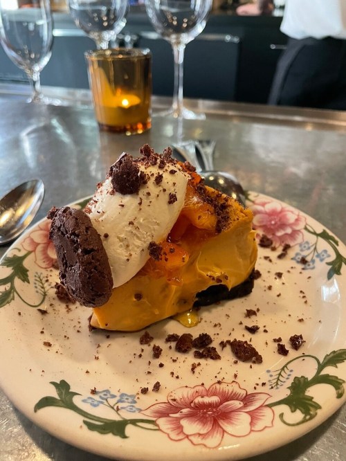 A slice of dessert on a floral plate, featuring layers of cream, chocolate cookie, and orange filling, garnished with whipped cream.