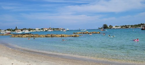 A serene beach scene with clear blue water, rocky outcrops, and people swimming in the distance under a sunny sky.