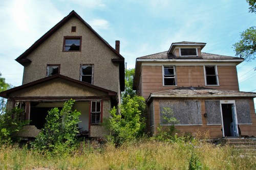 Two abandoned houses side by side, one with broken windows and overgrown grass, under a clear blue sky.
