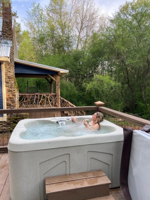 A person relaxes in a hot tub on a deck surrounded by trees and a rustic house in the background.