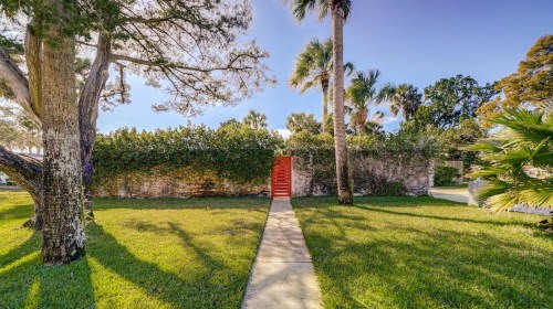 A vibrant red door framed by lush greenery and palm trees, with a stone pathway leading to it under a clear blue sky.