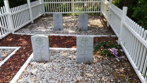 A small graveyard with four headstones surrounded by white fencing and decorative stones.