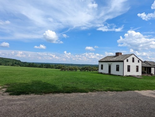 A white house sits on a grassy field under a blue sky with fluffy clouds, surrounded by rolling hills.