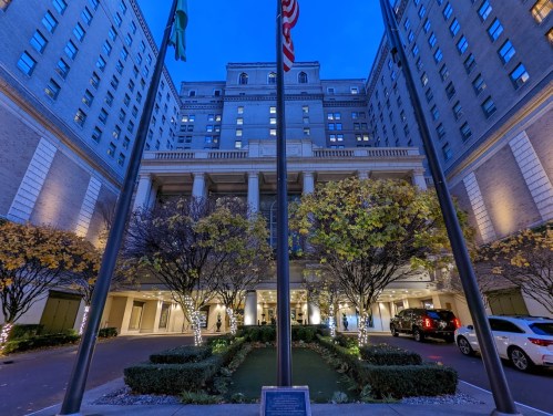 A grand hotel entrance at dusk, featuring flags, illuminated trees, and a landscaped courtyard.