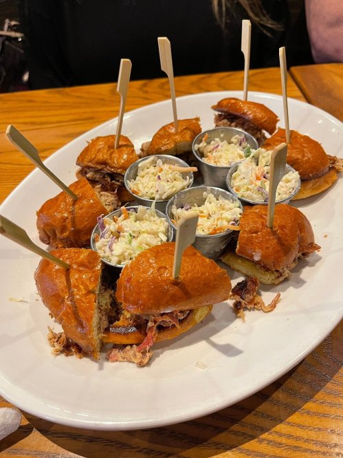 A platter of pulled pork sliders with coleslaw in small metal cups, garnished with wooden skewers.