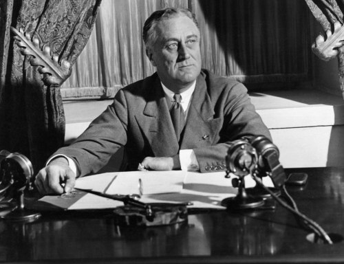 Black and white photo of a man in a suit sitting at a desk with microphones, looking thoughtfully at papers.