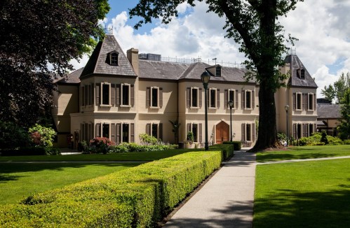 A large, elegant mansion with a manicured lawn and pathway, surrounded by trees and colorful landscaping.
