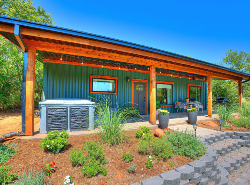 A modern blue house with a wooden porch, hot tub, and landscaped garden under a clear blue sky.