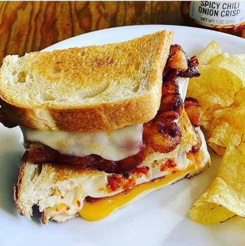 A stacked sandwich with bacon, cheese, and sauce, served with potato chips on the side.