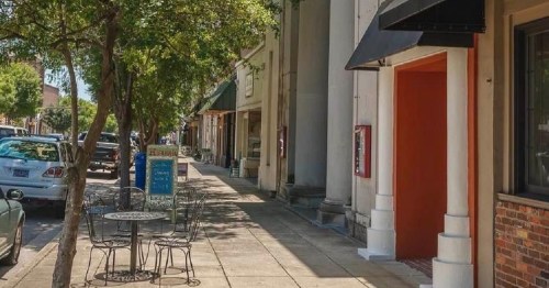A quiet street scene with outdoor seating, trees, and storefronts lining the sidewalk.