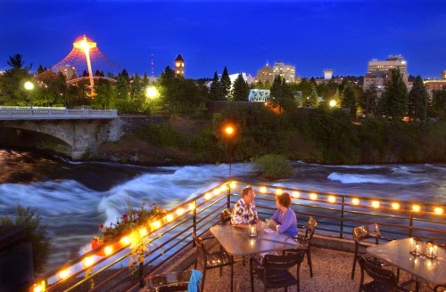 Couple dining on a riverside patio at night, with city lights and a bridge in the background.