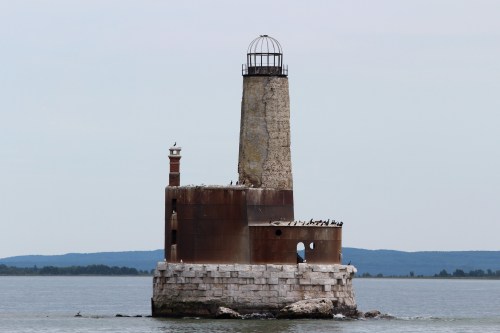 A weathered lighthouse stands on a rocky island, surrounded by calm water and distant hills under a clear sky.