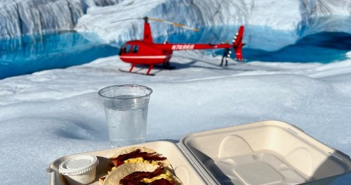 A meal of eggs and bacon on a snowy surface, with a helicopter and glacial water in the background.