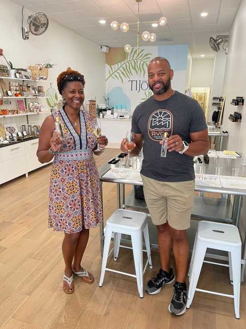 A man and woman hold small bottles in a bright store, smiling and standing by a table with beauty products.