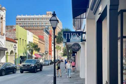 A vibrant street scene with shops, palm trees, and cars under a clear blue sky. People walk along the sidewalk.